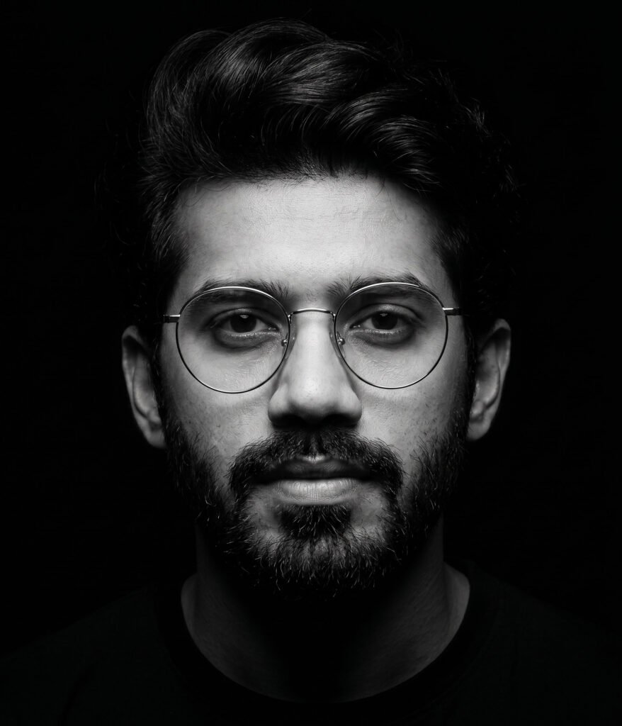 A black and white close-up portrait of a bearded man wearing round wire-rimmed glasses
